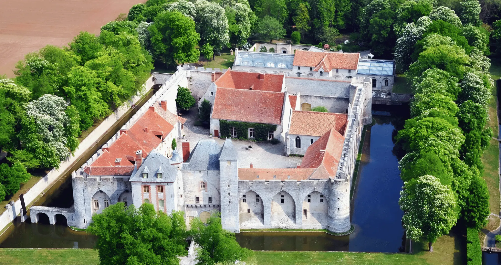 Le château dans ses douves vu du ciel : la cour, les toits, la verrière de la piscine derrière