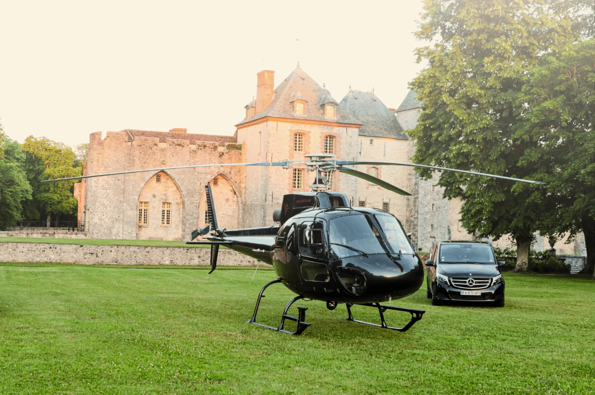 Un hélicoptère et deux voitures avec chauffeur sur la pelouse, le château au soleil du soir